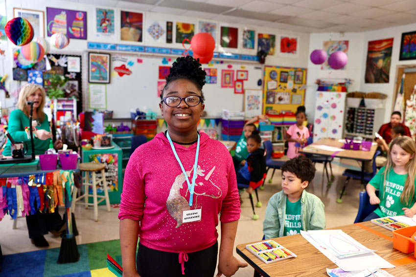 Smiling student standing in classroom with teacher and students in background
