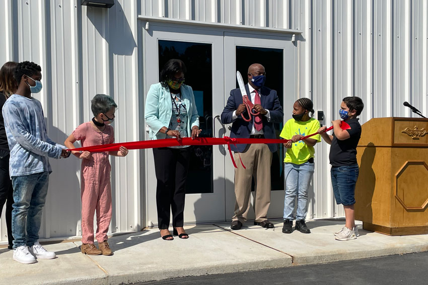 Administrators and students standing in front of new gym holding ribbon and oversized scissors